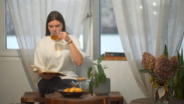 Young female student reads book in cafe. Media. Beautiful young woman comfortably spending time reading book in cafe. Cozy winter day in cafe with book and tea.