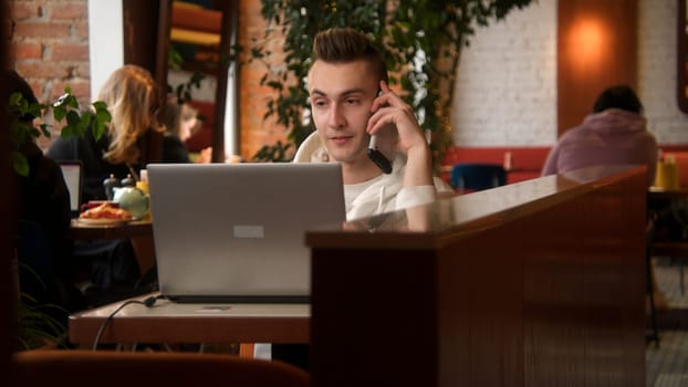 Young man is talking on phone at laptop in cafe. Stock footage. Young man is talking on phone while working on laptop. Man answers work calls while working on laptop in cafe.