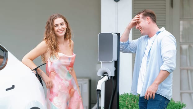 Happy and lovely couple with eco-friendly conscious recharging electric vehicle from EV home charging station. EV car technology utilized for residential home to future sustainability. Synchronos