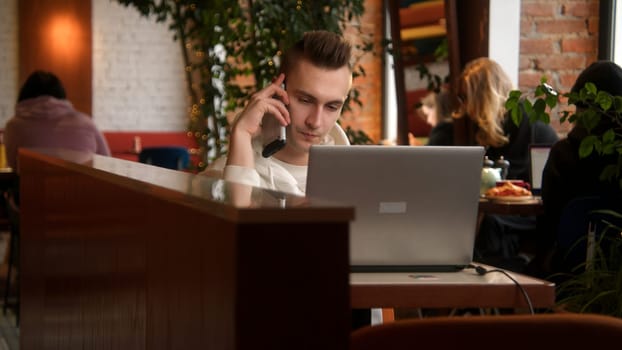 Young man is talking on phone on laptop. Stock footage. Handsome young man is talking on phone and working on laptop.