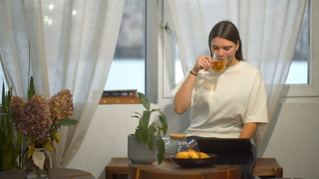 Young woman drinks tea and looks at tablet in cafe. Media. Student watches video on break and drinks tea in cozy cafe. Young woman is relaxing watching tablet and having tea in cafe.