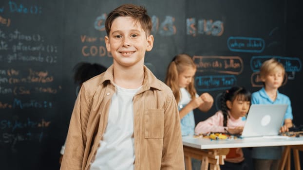 Smart boy smiling at camera while diverse friend working or learning engineering code or prompt in STEM technology classroom. Cute student standing at camera while children using laptop. Erudition.