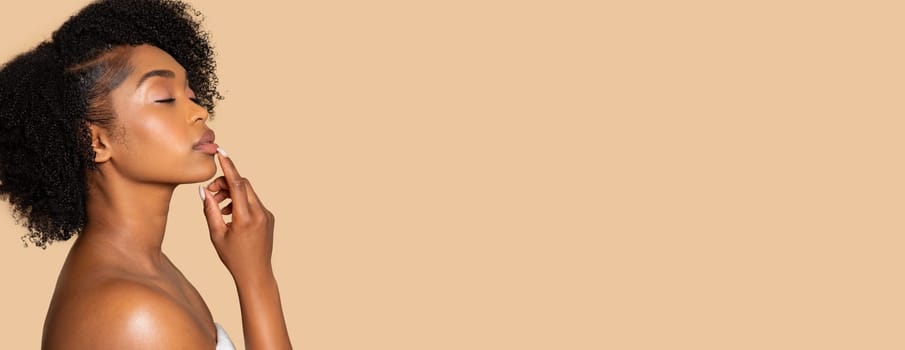 Pensive young black woman with curly hair, finger on her lips, demonstrating free space against spacious beige background, panorama, banner