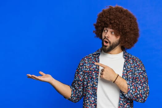 Happy amazed man with Afro hairstyle wig showing pointing left empty place, advertising area for commercial text copy-space for goods promotion advertisement logo text. Guy isolated on blue background