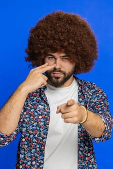 I am watching you. Confident attentive man pointing at his eyes and camera, show I am watching you gesture spying on someone. Stylish disappointed Caucasian guy isolated on blue background. Vertical