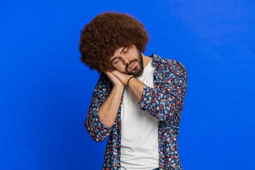 Tired exhausted young adult sleepy man with Afro hairstyle wig inattentive feeling somnolent lazy bored gaping suffering from lack of sleep, falling asleep. Guy isolated on studio blue background