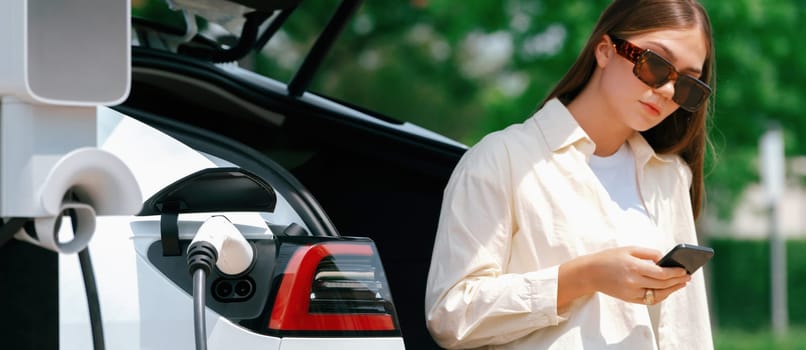 Young woman use smartphone to pay for electricity at public EV car charging station green city park. Modern environmental and sustainable urban lifestyle with EV vehicle. Panorama Expedient