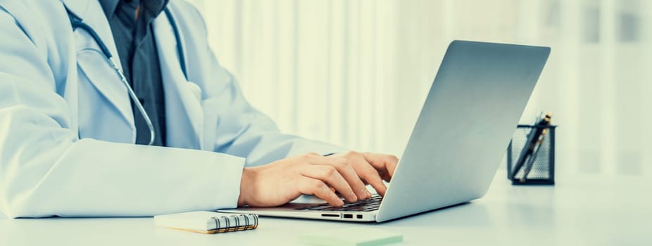 Doctor at hospital sit at his desk working on laptop diagnosing patient test results, developing treatment plan for illnesses and sicknesses. Medical staff and healthcare service. Panorama Rigid