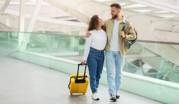 Trip Together. Happy European Couple Walking With Luggage At Airport Terminal, Romantic Young Man And Woman Embracing And Smiling To Each Other While Going To Boarding Gate, Copy Space