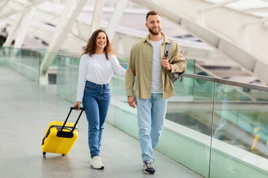 Relaxed young couple casually walking with luggage through the airport terminal, happy millennial spouses enjoying start of their vacation together, going to flight boarding gate, copy space