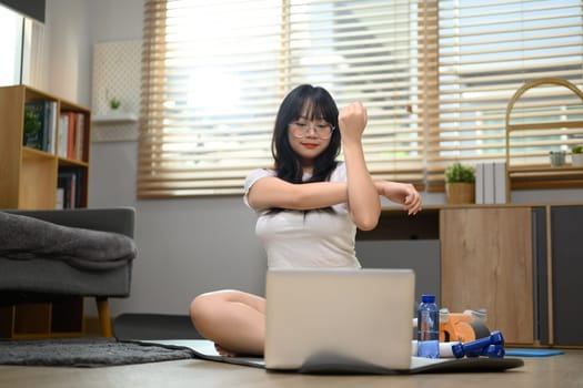 Smiling young woman stretching body on mat and watching online tutorial. Healthy lifestyle concept.