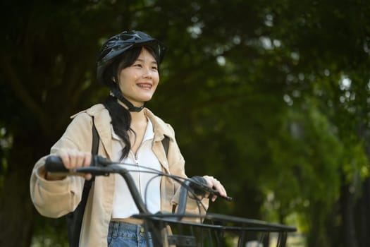 Smiling young woman wearing helmet walking in the park with her bicycle.