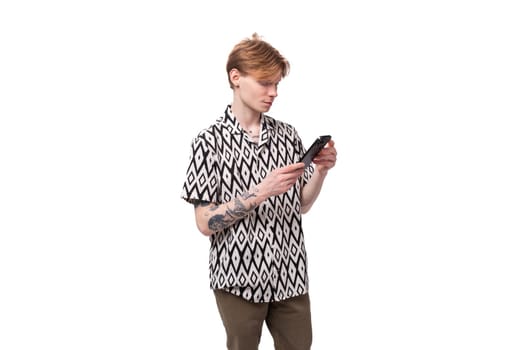 a young caucasian man with red hair and a tattoo on his arms is dressed in a stylish shirt chatting in a social network in a smartphone.
