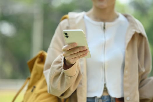 Young female student typing text on mobile phone while walking in the park.