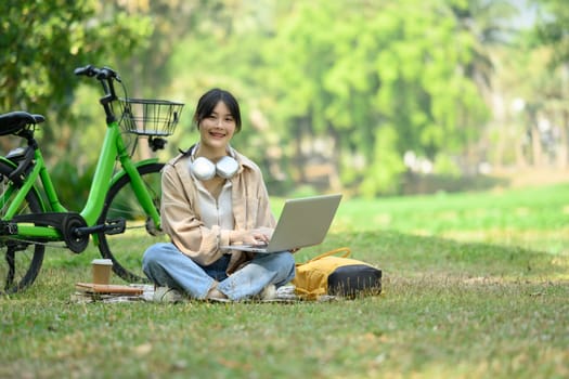 Full body shot of smiling Asian woman sitting on grass near bicycle and using laptop.