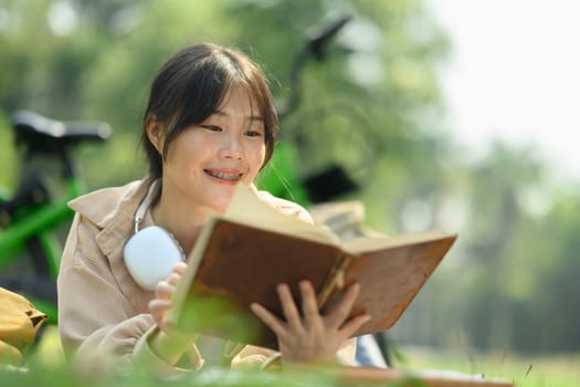 Delighted young Asian woman reading book on green grass during sunny day.