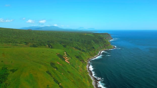 Wild nature, green mountains with steep green slopes leading to the sea. Clip. Aerial of foam of blue waves break in volcanic rocks