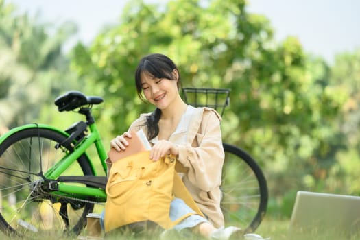 Positive young woman packing her backpack sitting on grass near her bicycle in the park.