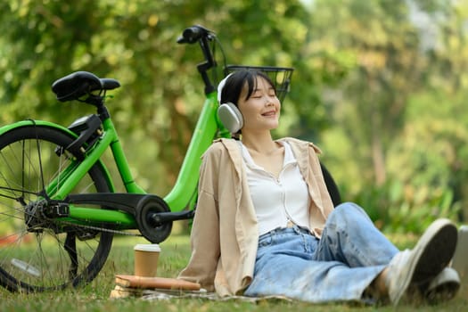 Happy young woman listening to music with headphone sitting at the park with her bicycle.