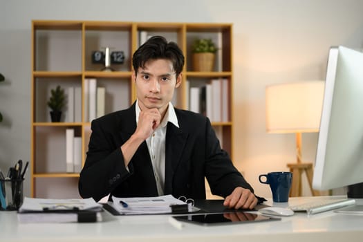 Confident millennial businessman sitting at workplace and looking at camera.