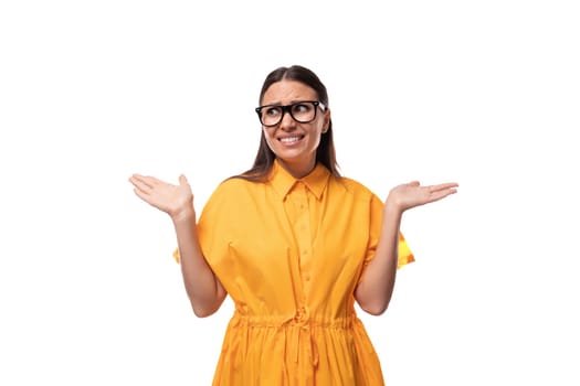 Caucasian young slim woman with black straight hair with glasses and in a yellow dress doubts on a white background.