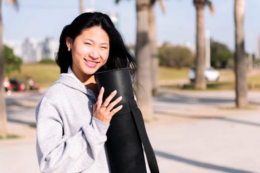 portrait of a young asian woman dressed in casual clothes smiling looking at camera while holding her yoga mat, healthy and active lifestyle concept, copy space for text