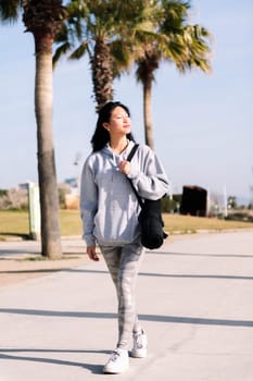 smiling young asian woman dressed in sportswear walking with yoga mat hanging on her arm, sport and healthy lifestyle concept