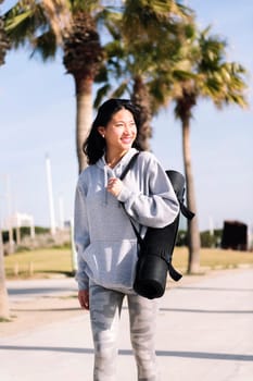 smiling young asian woman dressed in sportswear walking with yoga mat hanging on her arm, sport and healthy lifestyle concept