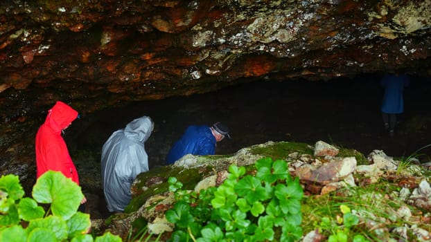 Group of tourists go to cave. Clip. Tourists enter cave in rocks on rainy day. Group of people in raincoats in rocky Mountains go to cave in summer.