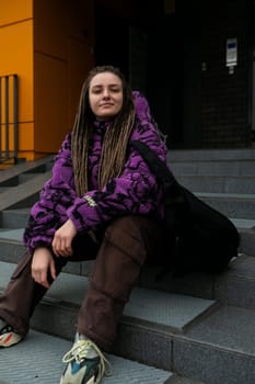 Lifestyle concept. Young woman with dreadlocks and piercing sits on the steps of a house.