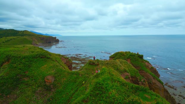 Top view of tourists on rocky coast trail with green grass. Clip. Beautiful landscape of rocky coast with green grass and tourists on hike. Tourists ride on edge of rocky coast overlooking sea on cloudy day.