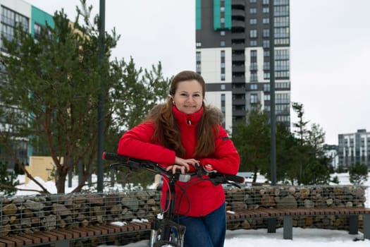 Pretty young woman in a winter jacket stands with a bicycle on the street.