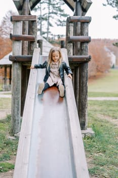 Little girl slides down a slide on a wooden playground holding the handrails. High quality photo