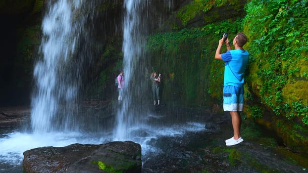 Tourists take pictures of waterfall on their phones. Clip. Tourists at water stream of rocky waterfall. Tourists at rocks with moss and waterfall in summer.