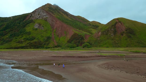 Top view of waves on coast with people and mountains. Clip. Trip to mountain coast of ocean in cloudy weather. People relax on north coast on backdrop of majestic mountains.