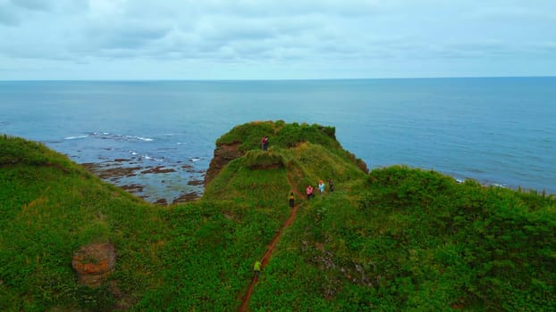 Top view of tourists on rocky coast trail with green grass. Clip. Beautiful landscape of rocky coast with green grass and tourists on hike. Tourists ride on edge of rocky coast overlooking sea on cloudy day.
