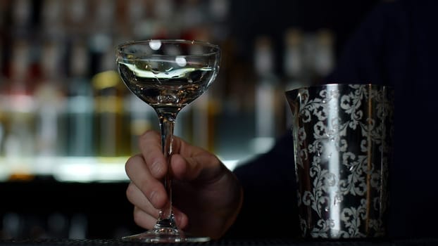 Close up of a bartender putting ice cubes into the small empty cocktail glass. Media. Details of making a drink with blurred bottles of alcohol on the background