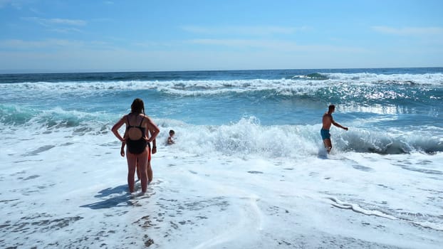 People bathe in beautiful blue waves on sunny day. Clip. Beautiful blue waves with foam on shore with vacationing people. People in swimsuits swim with blue waves of sea on summer day.