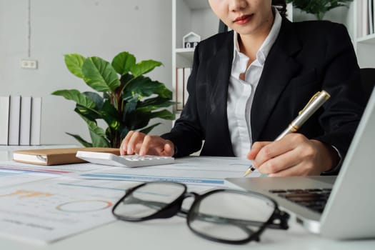 A woman is writing on a piece of paper with a pen. She is wearing a black suit and is sitting at a desk. The paper has a lot of numbers and calculations on it