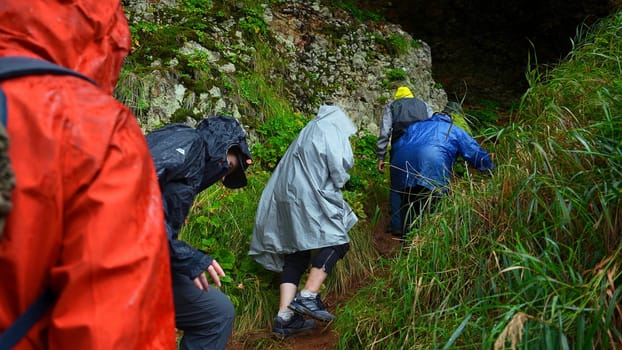 Group of tourists go to cave. Clip. Tourists enter cave in rocks on rainy day. Group of people in raincoats in rocky Mountains go to cave in summer.