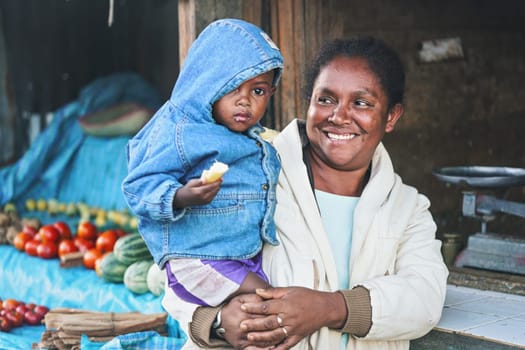 Ranohira, Madagascar - April 29, 2019: Unknown Malagasy woman holding her baby son on hands, smiling, blurred market stall background. People of Madagascar are poor but cheerful 