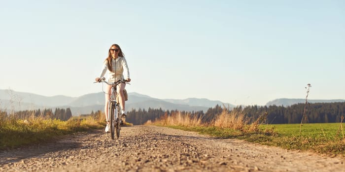 Young woman rides bicycle on dusty country road, smiling, blurred sunny countryside in background. Wide banner space for text right side