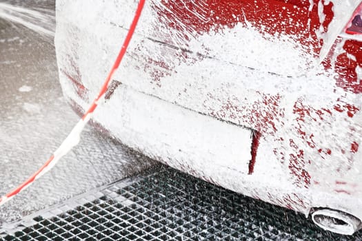 Red car washed in self serve carwash, detail on soap foam spraying from high pressure hose back, completely covered under white layer