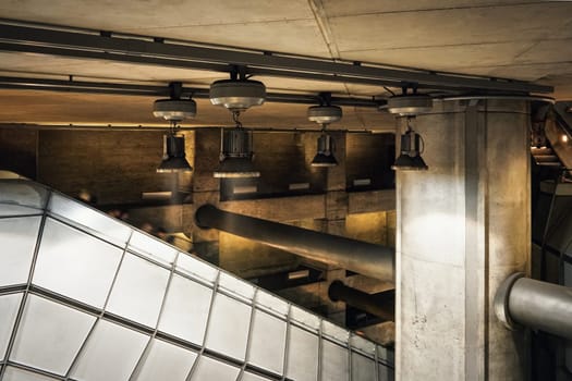 Industrial looking architecture, metal lamps shine over tubes, concrete walls and pillars in modern train station. Blurred people going down the escalator stairs