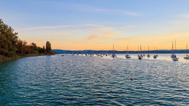 Bodensee Lake Sunrise Panorama. Morning Sunlight Over Tranquil Waters. Witness the mesmerizing dawn over Germany's Bodensee Lake, captured from a boat dock. Embrace the tranquil beauty of the early morning as the sun rises, casting a soft glow on the landscape. The peaceful scene features boats, yachts, and a charming water shack set against a backdrop of a captivating sky. Clouds delicately reflect on the calm water, creating a serene atmosphere. Immerse yourself in the serene beauty of a lakeside sunrise. Explore the harmony of nature, technology, and production as the day unfolds by the lake.