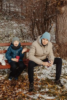 Capture the heartwarming essence of winter as two brothers, aged 8 and 17, share a special moment on a snow-covered bench in a serene rural park. Sipping hot tea from a thermos, they immerse themselves in the beauty of the snowy landscape, creating cherished memories of familial warmth amidst the tranquil winter scenery. This captivating image encapsulates the joy of family bonding, winter relaxation, and the simple pleasures of a cozy picnic in the midst of nature's snowy embrace.