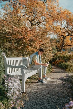 a teenager sits on a bench in the park drinks coffee from a thermo mug and looks into a phone. Portrait of handsome cheerful guy sitting on bench fresh air using device browsing media smm drinking latte urban outside outdoor.