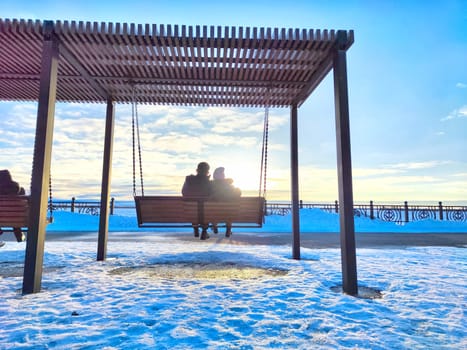 Two people sitting close on a swing under clear, cold sky in spring. Couple Enjoying a Winter Sunset on a Park Swing on the embankment