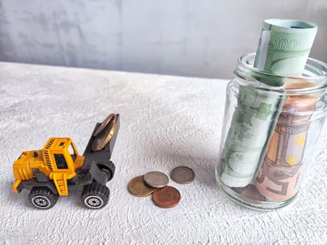 Toy Bulldozer Loading Coins Next to Jar of Cash. The concept of savings ranging from small money to large. The need for work to increase income