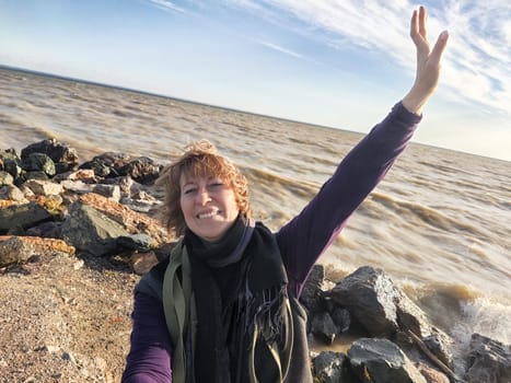 Happy cheerful middle aged woman in scarf taking selfie on nature outdoors and sun with water and waves of sea on the background